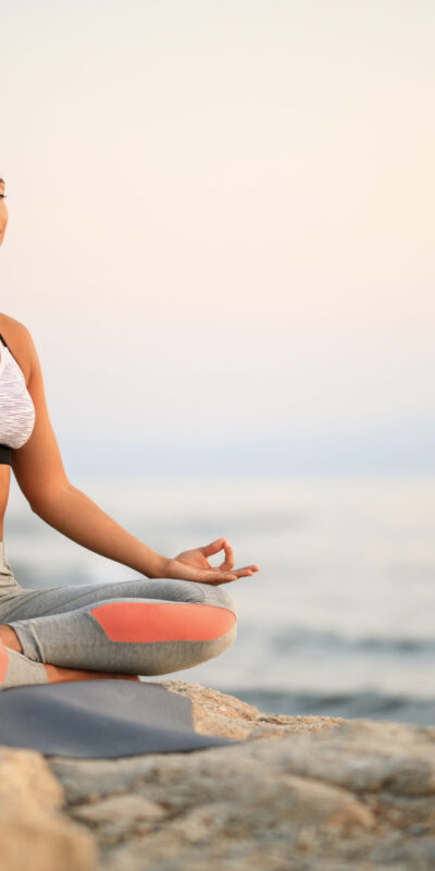 Young woman practicing Yoga relaxation exercises and meditating on a rock at the beach. Copy space.