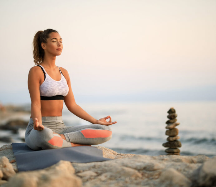 Young woman practicing Yoga relaxation exercises and meditating on a rock at the beach. Copy space.